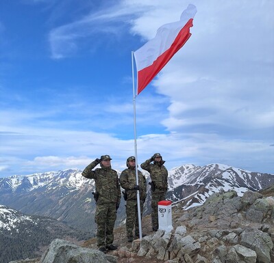 Funkcjonariusze straży granicznej oddają honor do Flagi Rzeczpospolitej polskiej na granicy w Tatarach. widoczne są szczyty gór a obok masztu z flagą znajduje się  słupek graniczny.