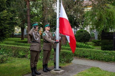 funkcjonariusze straży granicznej oddający honor przed masztem na który jeden z nich wciąga flagę Polski.