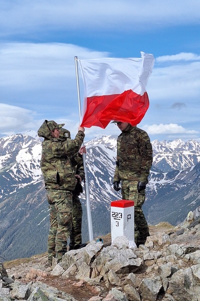 funkcjonariusze wieszają flagę Polski na maszt. uroczyste podniesienie flagi odbywa się na granicy ze słowacka w wysokich górach tatarach. wieje bardzo mocny wiatr. widoczne są szczyty górskie.