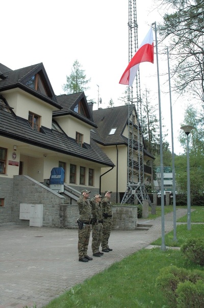 funkcjonariusze straży granicznej oddają honor przed flaga Polski. w tle widoczny jest budynek Placówki SG w Zakopanem.