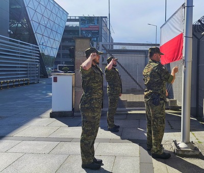 funkcjonariusze straży granicznej oddają honor przed flaga Polski. w tle budynek portu lotniczego w Krakowie-Balicach gdzie mieści się również Placówka SG..