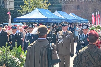 Na zdjęciu mężczyzna w mundurze SG stojący przodem. Po prawej i lewej stronie mężczyźni ubrani w mundury innych formacji. Na pierwszym planie mężczyzna w kapeluszu z piórkiem. W tle namioty wystawowe i tłum mieszkańców.