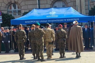Na zdjęciu grupa mężczyzn w umundurowaniu różnych formacji widoczna tyłem stoi przed namiotem wystawienniczym. W tle grupa innych mężczyzn widoczna przodem w mundurach oraz garniturach.
