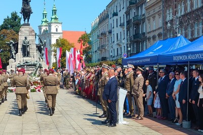 Po prawej stronie grupa mężczyzn i kobiet w garniturach, stojących pod namiotem wystawienniczym, przed nimi rząd mężczyzn w mundurach galowych różnych formacji. Po środku maszerują mężczyźni w mundurach. W tle widać pomnik.
