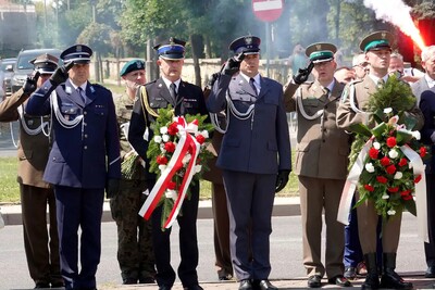 Na zdjęciu mężczyźni w umundurowaniu różnych formacji. Dwóch mężczyzn trzyma w rękach biało - czerwone wiązanki z kwiatów.