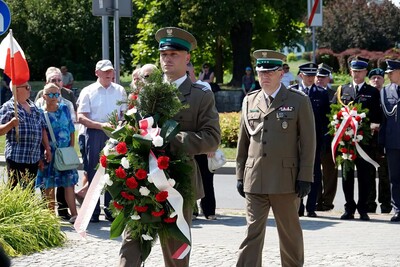 Na pierwszym planie mężczyzna w mundurze SG niosący w ręku wiązankę z biało - czerwonych kwiatów. Tuż za nim mężczyzna w mundurze SG. W tle grupa mężczyzn w umundurowaniu innych formacji, jeden z nich trzyma w ręku wiązankę z biało - czerwonych kwiatów. Z lewej strony grupa kobiet i mężczyzn, z których jeden trzyma w ręku biało - czerwoną flagę.