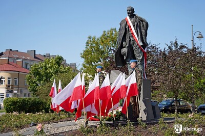 Na zdjęciu pomnik stojącego mężczyzny. Przed nim dwóch mężczy6zn w mundurach. Obok widoczne drzewa i pojazdy oraz zabudowania.