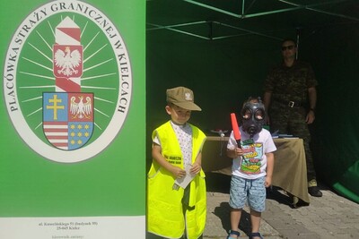 Na zdjęciu widoczna dwójka dzieci, jedno z nich trzyma latarkę do zatrzymywania pojazdów. W tle widoczna sylwetka funkcjonariusza, po lewej stronie baner reklamowy.