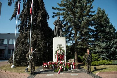 Na zdjęciu pomnik mężczyzny na postumencie, pod postumentem wiązanki i wieńce biało - czerwonych kwiatów. Obok pomnika dwie osoby, kobieta i mężczyzna w mundurach z bronią. W tle duże drzewa, po lewej stronie maszty z flagami państwowymi