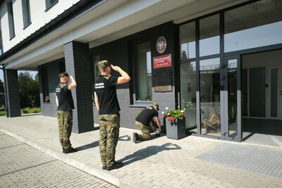 Na zdjęciu dwóch salutujących mężczyzn w umundurowaniu Straży Granicznej. Przed nimi mężczyzna kładący znicz przez tablicą pamiątkową wmurowaną w fasadę budynku.
