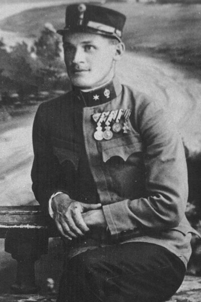Na zdjęciu mężczyzna w pozycji siedzącej z rękami opartymi o balustradę. Mężczyzna ubrany w historyczny mundur wojskowy, na głowie czapka wojskowa. W klapie marynarki munduru wpięte ordery.