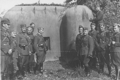 Na zdjęciu grupa mężczyzn w mundurach wojskowych z okresu II wojny światowej wraz z pasami, do których przypięta jest broń. Na głowach mężczyzn czapki wojskowe, część mężczyzn ma na rękach rękawiczki. Na środku schron bojowy z czasów II wojny światowej a przy nim mężczyzna także w mundurze wojskowym.