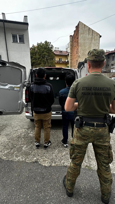 Na zdjęciu widać trzech mężczyzn. Jeden ubrany jest w mundur polowy przy pasie ma broń i stoi z dwoma mężczyznami, którzy mają wsiąść do samochodu.