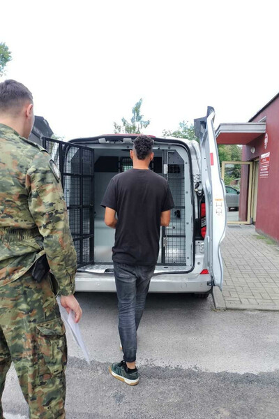 Na parkingu przed siedzibą placówki sg w kielcach stoi samochód służbowy sg typu więźniarka. przed wejściem do samochodu tyłem stoi zatrzymany cudzoziemiec, pilnowany przez funkcjonariusza sg