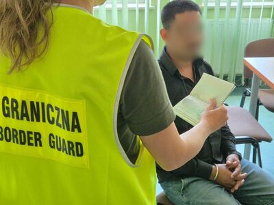 Na zdjęciu mężczyzna siedzący na krześle w pomieszczeniu służbowym placówki Straży Granicznej. Po lewej stronie kobieta w wyciągniętej ręce trzymająca dokument ubrana w kamizelkę odblaskową z napisem Straż Graniczna.