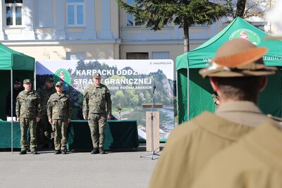 Zdjęcie przedstawia scenę plenerową podczas uroczystości pod gołym niebem, prawdopodobnie na placu lub przed budynkiem. Na pierwszym planie, z prawej strony, widoczna jest postać męska zwrócona plecami do obserwatora. Ma na sobie beżowy płaszcz i charakterystyczny kapelusz z rondem, ozdobiony piórkiem. Kapelusz jest brązowy z jasnym paskiem.
W centrum kadru, nieco dalej, stoi grupa pięciu mężczyzn w mundurach polowych w kamuflażu moro. Wszyscy są wyprostowani i stoją na baczność. Za nimi znajduje się duża, prostokątna plansza z napisem &quot;KARPACKI ODDZIAŁ STRAŻY GRANICZNEJ&quot;.
