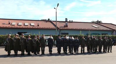 Zdjęcie przedstawia uroczysty apel na otwartym placu, prawdopodobnie przed budynkiem. W centrum kadru stoi długa, ustawiona w szeregu grupa młodzieży w mundurach. Od lewej strony, około ośmiu osób ubranych jest w ciemnobeżowe mundury, składające się z długich płaszczy, białych kołnierzyków, ciemnych spodni i kapeluszy z piórkami. Wszyscy mają założone białe rękawiczki. Za nimi, pośrodku szeregu, stoi kilku instruktorów lub dowódców w mundurach polowych w kamuflażu moro. Dalej, po prawej stronie, stoi grupa kilkunastu osób w ciemnych, szaro-granatowych mundurach w kamuflażu, z ciemnymi beretami i niebieskimi koszulami pod spodem. Po prawej stronie tej grupy stoi kolejna grupa w mundurach polowych w kamuflażu moro, również z ciemnymi beretami.