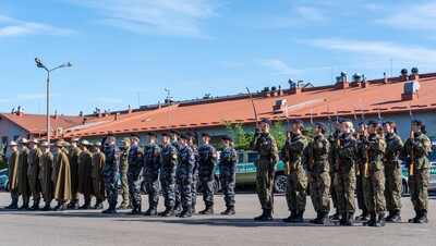 Zdjęcie przedstawia tę samą uroczystość co Obraz 2, ale z innej perspektywy. Długa grupa młodzieży w mundurach stoi na baczność. Od lewej strony, podobnie jak wcześniej, widoczne są osoby w ciemnobeżowych mundurach z długimi płaszczami i kapeluszami z piórkami. Dalej stoją osoby w ciemnych, szaro-granatowych mundurach w kamuflażu i ciemnych beretach. Następnie kolejna grupa w mundurach polowych w kamuflażu moro, również z ciemnymi beretami. Na prawym końcu szeregu grupa ta trzyma karabiny przewieszone przez ramię. Wszyscy są wyprostowani i patrzą w przód.