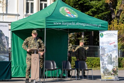 Zdjęcie przedstawia mężczyznę w mundurze polowym w kamuflażu moro, stojącego za drewnianą mównicą i wygłaszającego przemówienie. Mężczyzna jest w średnim wieku, ma krótkie włosy i patrzy w lewo. Na mównicy widoczne jest godło.
Za nim stoi zielony namiot z logo &quot;KARPACKI ODDZIAŁ STRAŻY GRANICZNEJ&quot; oraz adresem strony internetowej. Po prawej stronie namiotu ustawiono baner reklamowy z hasłem &quot;DOŁĄCZ DO NAS REALIZUJ PASJĘ MARZENIA&quot; i zdjęciami funkcjonariuszy Straży Granicznej w akcji.
Przed mównicą stoją cztery puste, czarne krzesła. W tle, po lewej stronie, widać fragment jasnego budynku z oknami, a po prawej – duże drzewo o zielonych liściach. Słońce świeci, a niebo jest jasne.