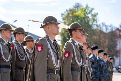 Zdjęcie przedstawia zbliżenie na grupę młodych mężczyzn w mundurach, ustawionych w szeregu. Są to prawdopodobnie absolwenci klas mundurowych. Na pierwszym planie widoczne są trzy postacie od pasa w górę. Wszyscy ubrani są w ciemnobeżowe mundury, składające się z marynarki z białymi ozdobami na ramionach i kołnierzu, białych koszul, krawatów oraz ciemnych kapeluszy z rondem, ozdobionych białym piórkiem. Na lewym ramieniu każdego munduru widoczna jest okrągła naszywka z polskim orłem i napisem &amp;quot;STRAŻ GRANICZNA&amp;quot;. Mężczyźni patrzą w prawo, a ich twarze są poważne i skupione.