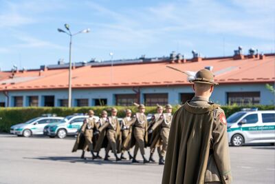 Zdjęcie przedstawia plecy osoby w beżowym płaszczu i kapeluszu z piórkiem, stojącej na pierwszym planie i patrzącej w kierunku grupy maszerujących osób. W tle, w oddali, widoczna jest kolumna ośmiu osób w ciemnobeżowych mundurach z długimi płaszczami i kapeluszami z piórkami, maszerujących w równym szeregu.
Za maszerującymi osobami widać kilka samochodów Straży Granicznej – biało-zielone furgonetki i osobowe pojazdy. W tle rozciąga się długi, niski budynek z pomarańczowym dachem.