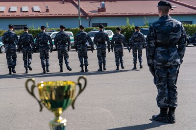 Zdjęcie przedstawia z perspektywy niskiego kąta trofeum sportowe na pierwszym planie oraz grupę uczniów w mundurach w tle. Na pierwszym planie, lekko rozmyty, stoi złocisty puchar z zielonymi akcentami i rzeźbionymi uchwytami.
W tle, nieco dalej, widoczna jest grupa uczniów w ciemnych, szaro-granatowych mundurach w kamuflażu, z ciemnymi beretami. Uczniowie stoją w równym szeregu, zwrócone plecami do obserwatora, patrząc w kierunku osoby stojącej po prawej stronie kadru, również w tym samym mundurze, ale zwróconej przodem do szeregu.
Za uczniami widać szereg zaparkowanych biało-zielonych samochodów Straży Granicznej. W tle rozpościera się długi, niski budynek z pomarańczowym dachem. Nad budynkiem widać błękitne niebo z białymi chmurami.