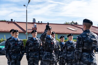 Zdjęcie przedstawia uroczystość ślubowania lub apelu. Na pierwszym planie, od prawej strony, stoi młody mężczyzna w ciemnym, szaro-granatowym mundurze w kamuflażu i ciemnym berecie, zwrócony przodem do grupy uczniów.
Przed nim, po lewej stronie, stoi szereg młodych mężczyzn i kobiet w tych samych mundurach i beretach. Mają na sobie niebieskie koszule pod mundurem. Jeden z nich, stojący w środku szeregu, podnosi prawą rękę w geście przysięgi lub meldunku. Inni stoją na baczność, patrząc w kierunku osoby dowodzącej.
W tle widoczny jest długi, niski budynek z pomarańczowym dachem oraz zaparkowane samochody Straży Granicznej. Niebo jest błękitne z delikatnymi chmurami.