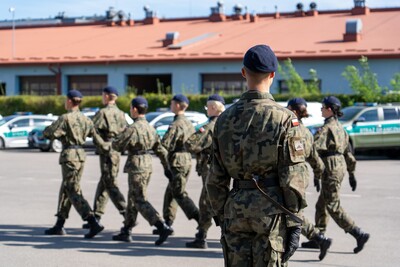 Zdjęcie przedstawia grupę młodych ludzi w mundurach polowych w kamuflażu moro, z ciemnymi beretami, maszerujących w równym szeregu, od lewej do prawej strony kadru. Wszyscy są zwróceni profilem do obserwatora.
Na pierwszym planie, po prawej stronie, stoi osoba w takim samym mundurze, zwrócona plecami do obserwatora, z rękami opuszczonymi wzdłuż ciała. Na jej plecach widoczny jest mały, czarny kabelek, prawdopodobnie od krótkofalówki lub innego sprzętu komunikacyjnego. Na ramieniu munduru widoczna jest naszywka z polskim orłem i napisem.
W tle za maszerującymi osobami widoczne są zaparkowane biało-zielone samochody Straży Granicznej. Dalej w tle rozpościera się długi, niski budynek z pomarańczowym dachem. Niebo jest błękitne z niewielkimi chmurami.