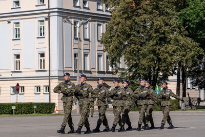 Zdjęcie przedstawia grupę siedmiu młodych ludzi w mundurach polowych w kamuflażu moro, z ciemnymi, niebieskimi beretami, maszerujących w równym szeregu przez duży, otwarty plac. Uczniowie maszerują od lewej do prawej strony kadru, z wyprostowanymi sylwetkami i jednolitym krokiem. Na ich koszulach pod mundurem widać niebieskie kołnierzyki.
W tle po lewej stronie widać fragment dużego, jasnego, klasycznego budynku z wieloma oknami. Pośrodku tła rośnie duże, zielone drzewo. Po lewej stronie kadru widać znak &quot;STOP&quot; i fragment zielonego żywopłotu. Niebo jest błękitne z kilkoma białymi chmurami.