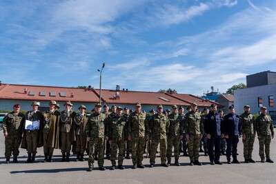 Przedstawia grupę około 15-20 mężczyzn, ułożonych w dwa rzędy. W pierwszym rzędzie, po lewej stronie, stoi dwóch mężczyzn w mundurach polowych, uściskiem dłoni gratulują sobie nawzajem. Jeden z nich, z beretem w kolorze bordowym, wręcza drugiemu dyplom. Za nimi stoi grupa młodych ludzi w jasnobrązowych, historycznych mundurach wojskowych, przypominających te z czasów przedwojennych. Mają na głowach brązowe kapelusze. Trzymają dwa puchary. Po prawej stronie zdjęcia stoją żołnierze w mundurach polowych oraz w granatowych beretach, które wyróżniają się na tle pozostałych. Wszyscy mężczyźni są wyprostowani, patrzą w kierunku aparatu.
