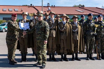 Zdjęcie przedstawia dużą grupę około 25 mężczyzn stojących w rzędzie na utwardzonym placu. W tle widoczny jest długi, parterowy budynek z czerwonym dachem oraz fragmenty innych, nowoczesnych budynków. Po lewej stronie grupy stoi kilku młodych ludzi w jasnobrązowych, historycznych mundurach z kapeluszami. Pomiędzy nimi a pozostałymi, w centralnej części zdjęcia, stoją starsi mężczyźni w mundurach polowych. Po prawej stronie grupy, w tylnym rzędzie, stoi kilku mężczyzn w granatowych beretach i ciemnych mundurach. Wszyscy patrzą w kierunku obiektywu, a za nimi rozpościera się jasne, błękitne niebo z kilkoma białymi chmurami.