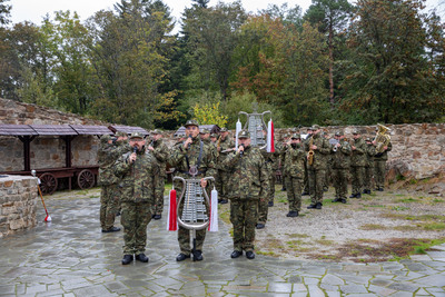 Wojskowa orkiestra dęta w mundurach maskujących stoi w szyku na kamiennym dziedzińcu. Dwa rzędy żołnierzy trzymają instrumenty, a dwóch na pierwszym planie ma mikrofony i trzyma ozdobną lirę orkiestrową. W tle kamienne mury i las.