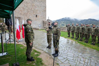 Dwóch funkcjonariuszy Straży Granicznej w mundurach wojskowych stoi frontem do siebie, wykonując komendy na placu obok kamiennej ściany. W tle, na błyszczącej od deszczu nawierzchni, stoi w szyku oddział żołnierzy. Dalej widoczne zalesione, zamglone wzgórza.
