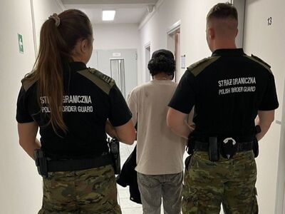 Dwóch funkcjonariuszy Straży Granicznej w mundurach prowadzi korytarzem budynku cudzoziemca ubranego w jasny T-shirt i spodnie jeansowe.