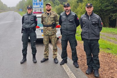 Czterech funkcjonariuszy słowackiej Policji, w tym jeden w mundurze moro, stojących w pełnym umundurowaniu obok radiowozu na granicy, na tle znaków drogowych.