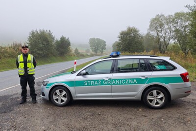 Funkcjonariusz polskiej Straży Granicznej w kamizelce odblaskowej stojący obok srebrnej Skody Octavii z zielonymi pasami i napisem „STRAŻ GRANICZNA” na tle mglistego, wiejskiego krajobrazu.