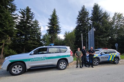 Trzech funkcjonariuszy Straży Granicznej i Policji stojących między samochodem terenowym Straży Granicznej (Toyota) a radiowozem słowackiej Policji, na tle wysokich świerków i słonecznego nieba.
