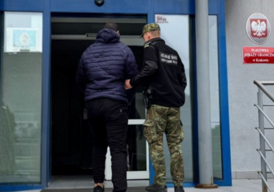 Mężczyzna w mundurze prowadzi do budynku osobę zatrzymaną. Ubrany jest w granatową kurtkę i buty sportowe. Wchodzą po schodach do budynku. Z prawej strony zdjęcia widać czerwoną tabliczkę z napisem Placówka Straży Granicznej w Krakowie.