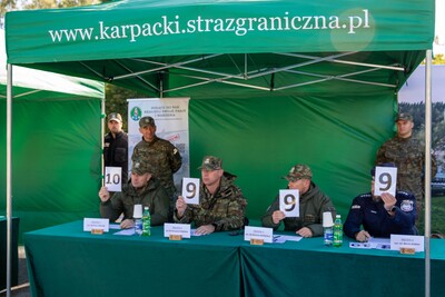 Czterech mężczyzn w mundurach siedzi za zielonym stołem pod zielonym namiotem z napisem &#039;www.karpacki.strazgraniczna.pl&#039;. Trzech z nich trzyma tabliczki z cyframi &#039;9&#039; i &#039;10&#039;.&quot;