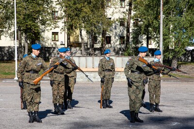 Grupa młodych żołnierzy w mundurach polskiej armii i niebieskich beretach stoi na zewnątrz, trzymając karabiny. W tle budynki i drzewa.