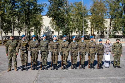 Grupa dziesięciu żołnierzy w mundurach, w tym ośmiu w niebieskich beretach, stoi w szyku razem z kobietą w jasnym płaszczu. Za nimi drzewa i budynek.