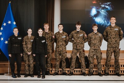 ośmioro uczniów - 3 kobiety i 5 mężczyzn - w umundurowaniu stoi przy fladze UE