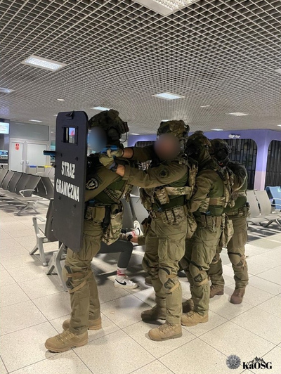 Funkcjonariusze ZIS w oporządzeniu  bojowym z tarczą balistyczna poruszają się na terenie terminala w zwartej grupie szturmowej