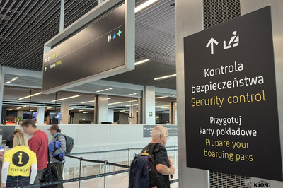 Na zdjęciu widać halę na lotnisku. Po prawej stronie zdjęcia widać tablicę z napisem kontrola bezpieczeństwa. Po lewej stronie zdjęcia widać osoby z bagażami, które przygotowują się do kontroli.