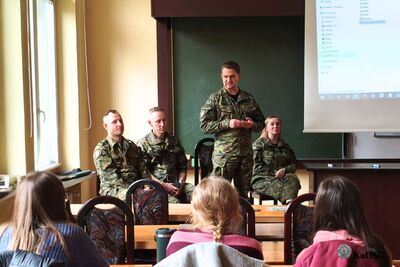 Mężczyzna w mundurze Straży Granicznej stoi i przemawia do słuchaczy, podczas gdy trzech oficerów (dwóch mężczyzn i jedna kobieta) siedzi obok. Na ekranie widoczny fragment listy lub struktury danych.