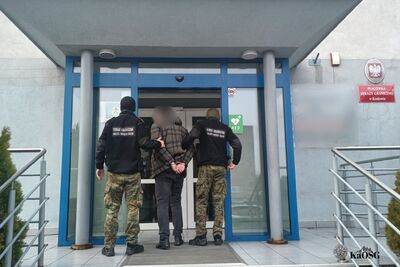 Na zdjęciu mężczyzna prowadzony przez dwóch mundurowych najprawdopodobniej funkcjonariuszy Straży Granicznej. Z tyłu na rękach ma kajdanki. W tle budynek z godłem na ścianie i tabliczką informującą o siedzibie Placówki SG w Krakowie.