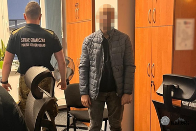 Zdjęcie zrobione we wnętrzu, prawdopodobnie w biurze Straży Granicznej, z widocznymi drewnianymi szafami na środku. Na pierwszym planie po prawej stoi młody mężczyzna w szarej, pikowanej kurtce i ciemnych spodniach. Jego twarz jest ocenzurowana (rozpikselowana). Po lewej stronie, tyłem do obiektywu, stoi funkcjonariusz Straży Granicznej w ciemnym mundurze z krótkim rękawem. Na plecach munduru widnieje napis: &quot;STRAŻ GRANICZNA POLISH BORDER GUARD&quot;. W pomieszczeniu znajdują się również ciemne fotele biurowe.