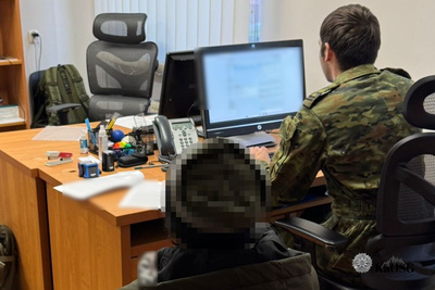 Zdjęcie zrobione we wnętrzu, prawdopodobnie w biurze. Na pierwszym planie widoczne jest duże, jasne drewniane biurko, na którym leżą dokumenty, telefon stacjonarny i akcesoria biurowe. Po prawej stronie, tyłem do obiektywu, siedzi funkcjonariusz w mundurze w kamuflażu (oliwkowo-brązowym) i pracuje przy monitorze komputera. Obok monitora widać drugi, wyłączony ekran. Przy biurku, bliżej obiektywu, siedzi inna osoba (jej głowa jest ocenzurowana, zakryta pikselami), widoczna głównie od tyłu.