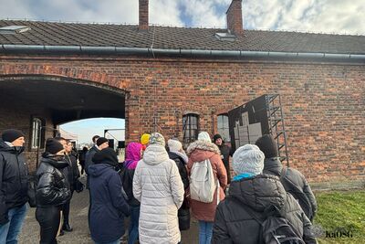 Grupa osób w kurtkach zimowych stoi przed ceglanym budynkiem z dużym przejściem łukowym (bramą) i dwoma kominami na dachu. W tle widać fragment nieba i namiot. Osoby patrzą na tablicę po prawej stronie, a jedna z nich zdaje się prowadzić prelekcję.