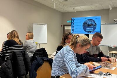 Kilka osób siedzi przy stołach w nowoczesnym pomieszczeniu, biorąc udział w zajęciach edukacyjnych. Na ścianie po prawej stronie znajduje się duży monitor z wyświetloną grafiką i tekstem, częściowo zasłoniętą. Ludzie koncentrują się na materiałach leżących na stołach i tabletach.